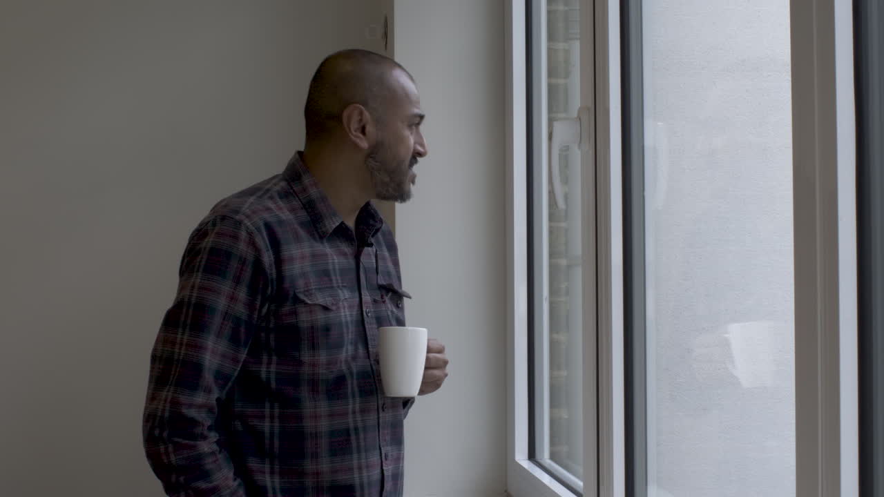 Indian Male Walking Towards Large Windows Holding White Mug Looking Out Contemplatively