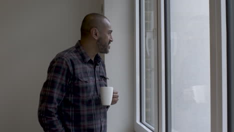 indian male walking towards large windows holding white mug looking out contemplatively