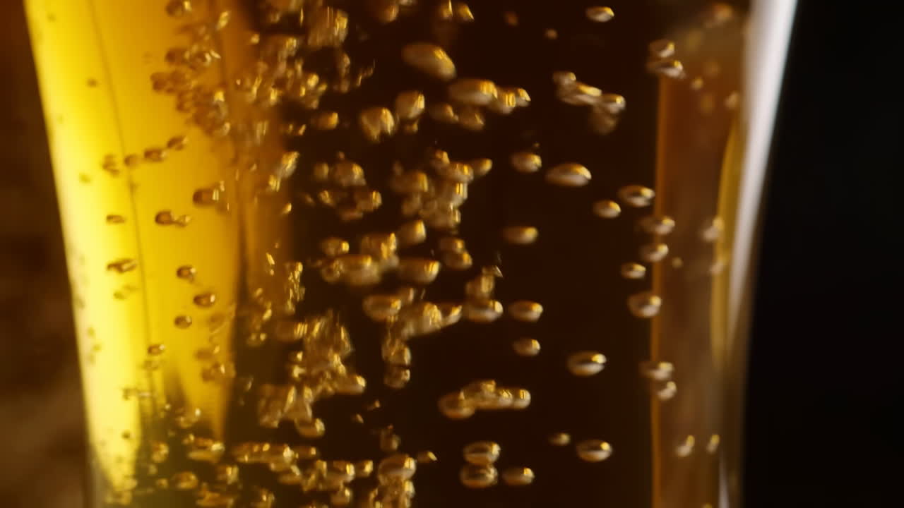 Primo piano di bicchiere di birra ambrata con bollicine che salgono, ripresa cinematografica al rallentatore di carbonazione dorata, luce