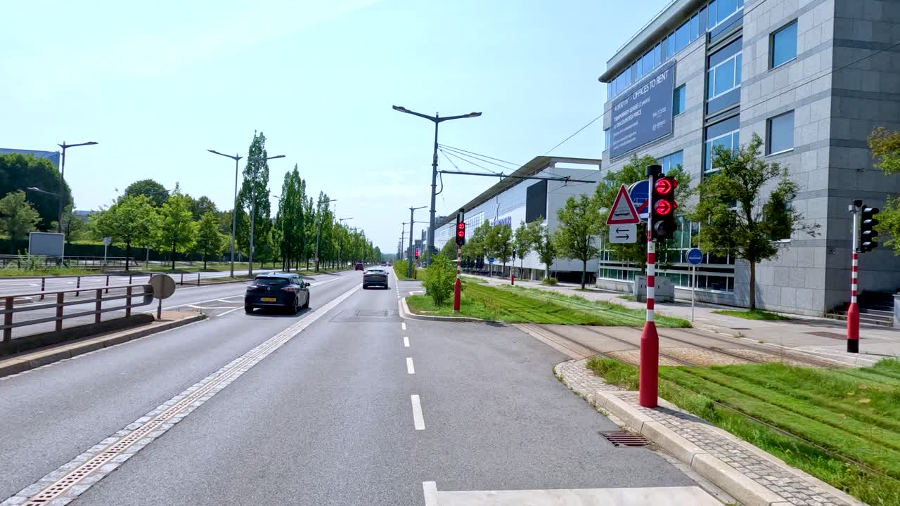 A black car travels down a sunlit urban street lined with modern glass buildings, greenery, and tram lines in Luxembourg. Smooth camera movement, bright daylight