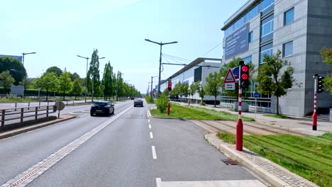 a black car travels down a sunlit urban street lined with modern glass buildings, greenery, and tram lines in luxembourg. smooth camera movement, bright daylight