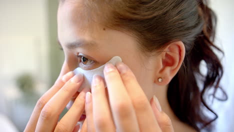 Portrait-of-smiling-biracial-teenage-girl-putting-on-under-eye-mask-in-bathroom,-slow-motion