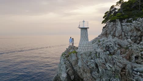 Isla-De-Kalamota,-Mar-Adriático,-Croacia---Una-Pareja-Saboreando-La-Puesta-De-Sol-Junto-Al-Faro---Toma-Aérea-De-Retroceso