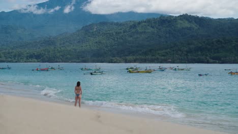 high quality cinematic shot of a woman standing on a remote tropical island beach with a volcano and fishing boats in the background and crystal clear blue water
