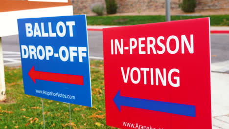 blue ellection ballot drop-off sign with red in-person voting sign blowing in the wind outside