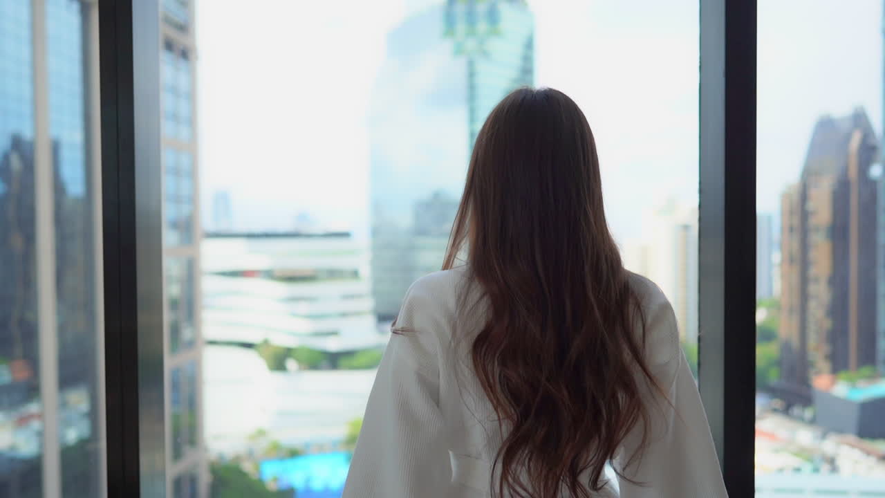 A woman with long brunette hair and her back to the camera stretches while looks out over the skyline of Bangkok from her hotel window