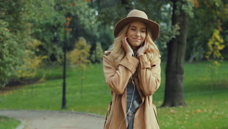 mujer rubia joven caucásica poniéndose los auriculares y escuchando la música mientras camina en el parque en otoño