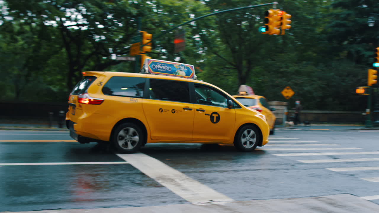 New York, USA, 25. maj 2025: Gul New York-taxa kører på våd bygade. Sidebillede af gul minivan-taxa, der kører gennem Manhattan-kryds med træer og trafiklys i baggrunden