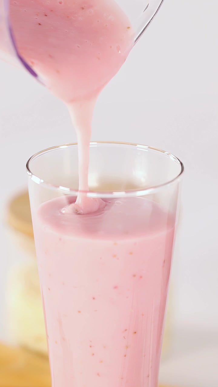 Un frullato cremoso rosa alla fragola si versa delicatamente in un bicchiere trasparente, con illuminazione brillante e vista ravvicinata.