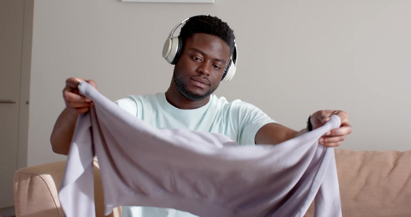 African American man holding sweatshirt and inspecting on sofa at home, wearing headphones, folding