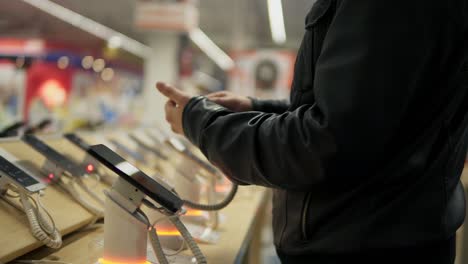 Young-male-customer-choosing-a-new-mobile-phone-in-a-shop.-He-is-trying-how-it-works