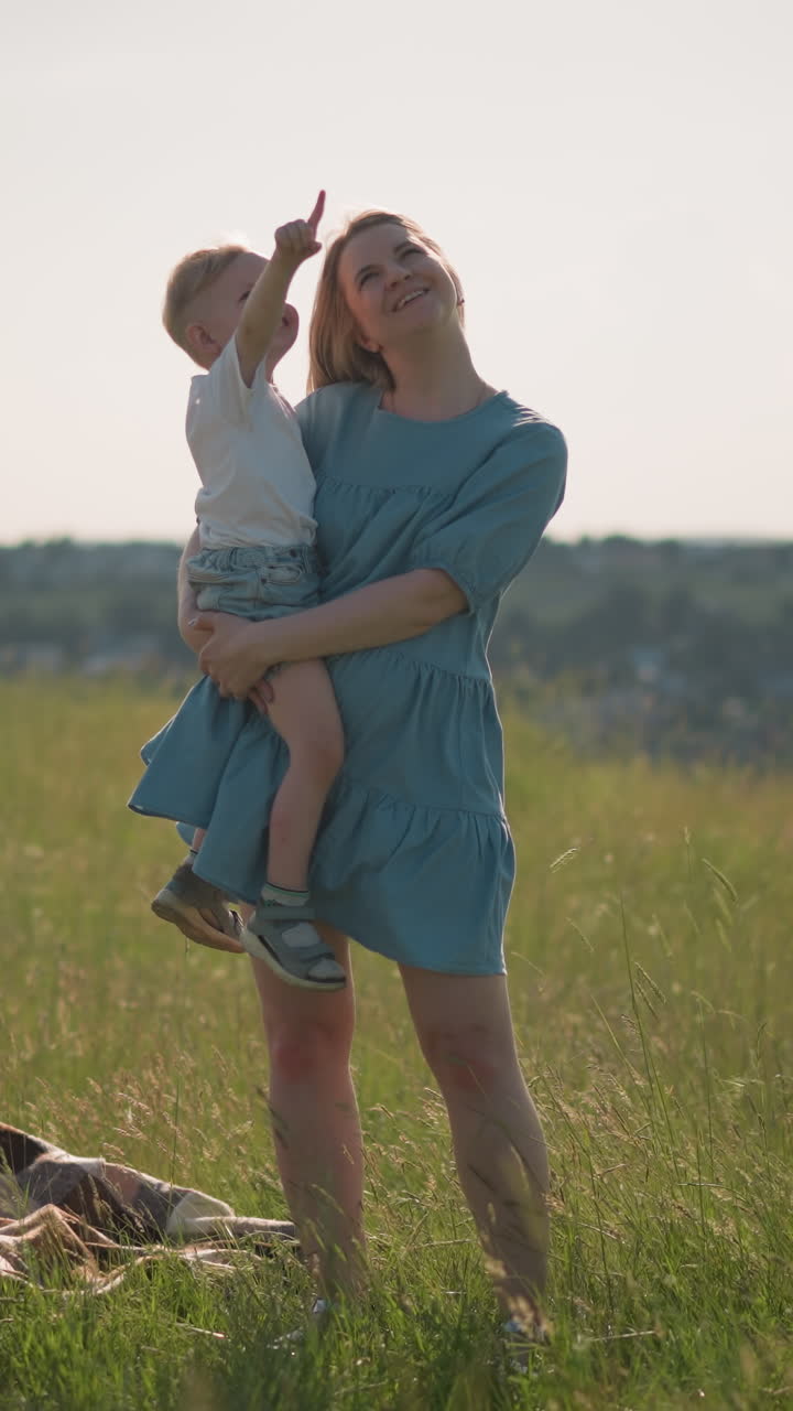una mujer con un vestido azul lleva a su hijo pequeño, que está señalando a los pájaros, mientras que su hermano está de pie en el fondo. ambos niños llevan camisetas blancas. una bufanda a cuadros se encuentra en la hierba