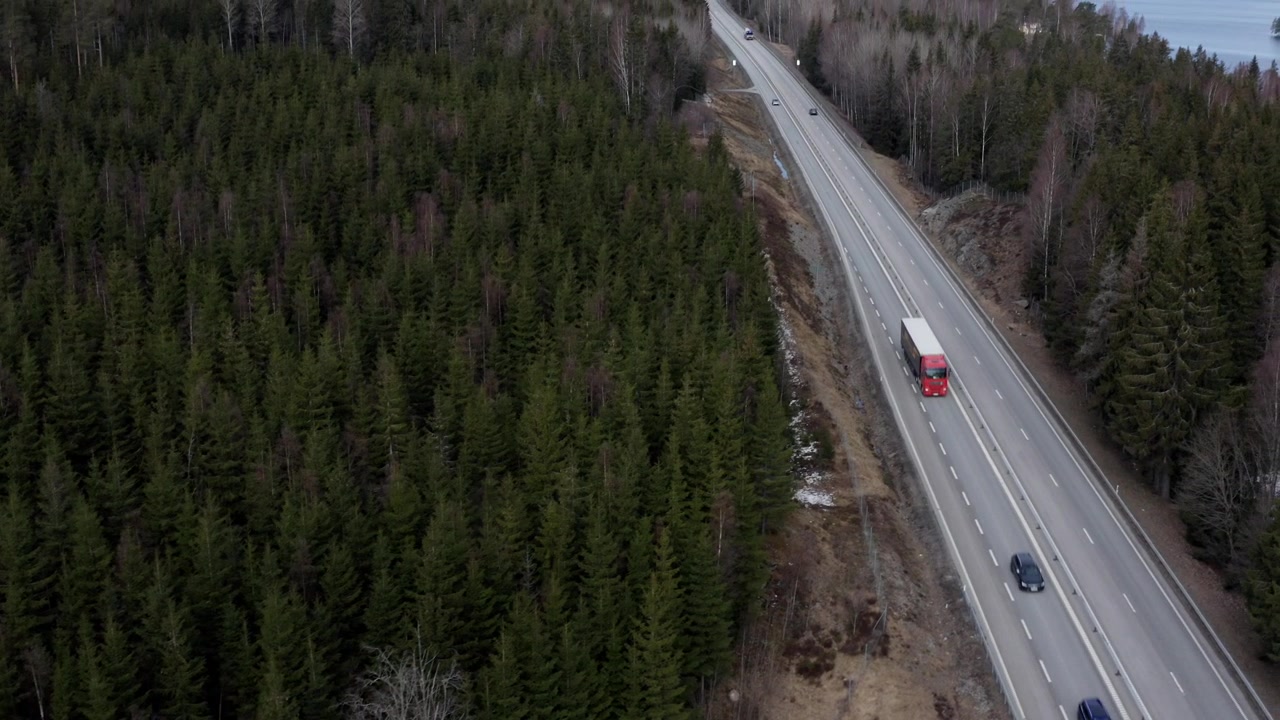 Ripresa con drone di un'autostrada in Svezia che attraversa una foresta, con panoramica verso l'alto per rivelare camion e auto in transito, un lago e il cielo.
