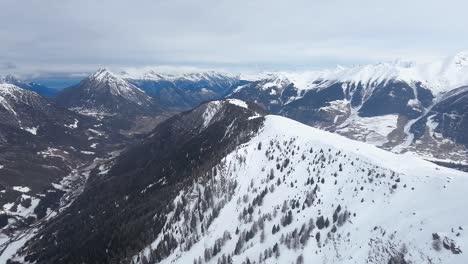 A-breathtaking-aerial-view-of-a-snow-covered-mountain-ridge-with-forested-slopes,-surrounded-by-a-majestic-range-of-peaks-under-a-cloudy-sky