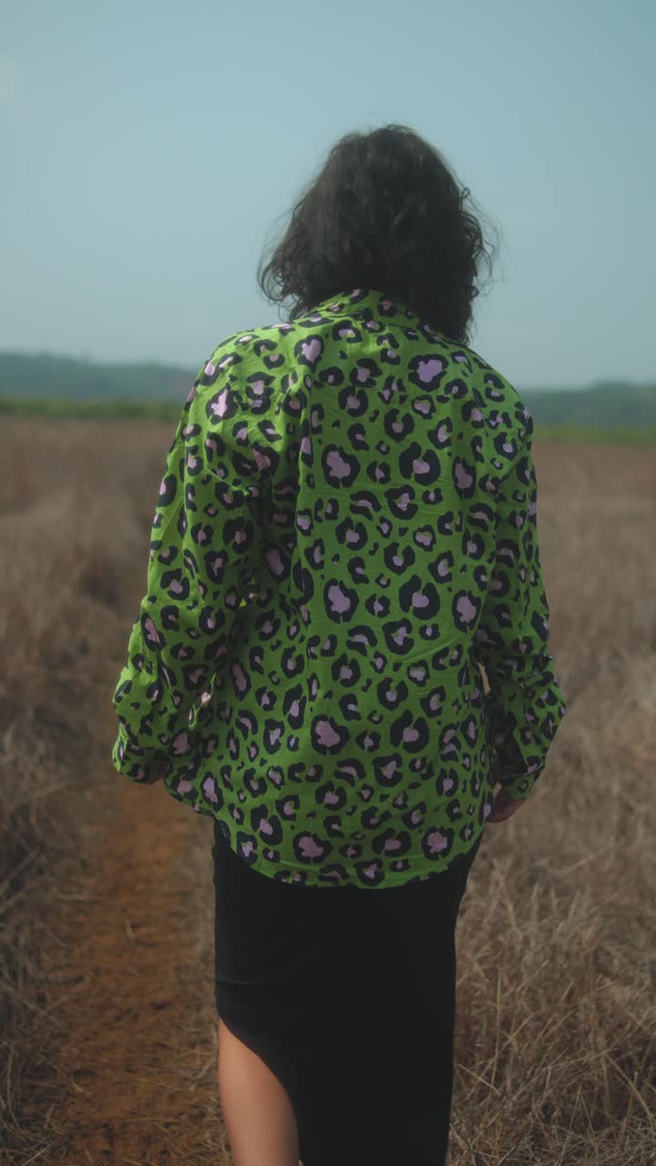 Woman walks alone on dry grassy path, touching her hair under clear blue sky