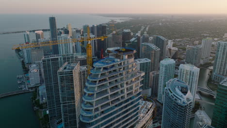 Aerial-cinematic-shot-of-top-of-skyscraper-under-construction.-Crane-towering-above-modern-development.-Futuristic-urban-borough-on-waterfront.-Miami,-USA