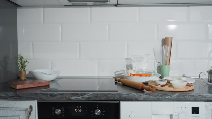 Bellissima cucina moderna con elegante piano di lavoro in marmo nero, pensili bianchi e forno incorporato. Utensili da cucina, matterello, bollitore in vetro e ingredienti per dolci disposti ordinatamente sull'area di lavoro