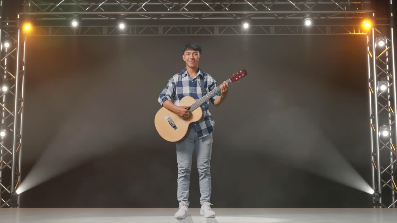 cuerpo lleno de adolescente asiático con guitarra sonriendo mientras está de pie en el escenario