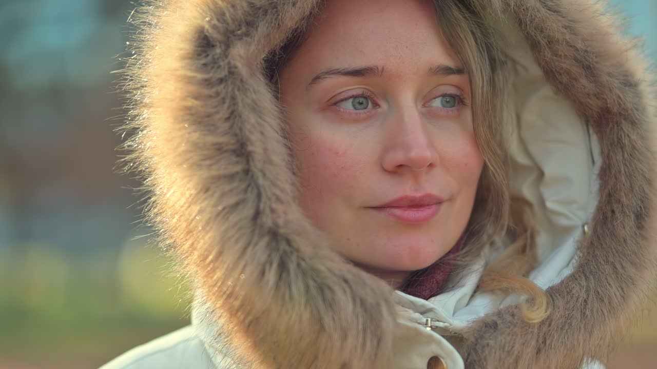 A woman walks through a peaceful park on a cool winter day. She wears a cozy fur-lined coat and smiles as she enjoys the beautiful surroundings filled with trees and natural light
