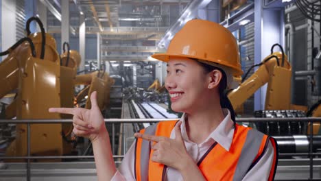 close up of asian female engineer with safety helmet smiling and pointing to side while standing with industrial robotic arms working