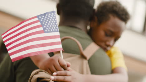 porträt eines traurigen jungen, der eine usa-flagge hält und seinen unerkennbaren militärvater umarmt