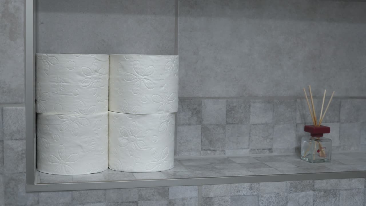 A person’s hand reaching for a roll of toilet paper on a bathroom shelf. The neatly stacked toilet paper rolls contrast with the modern tiled background, creating a clean and organized appearance.