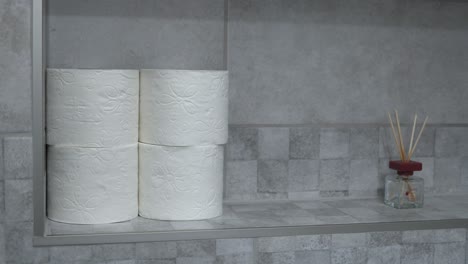 a person’s hand reaching for a roll of toilet paper on a bathroom shelf. the neatly stacked toilet paper rolls contrast with the modern tiled background, creating a clean and organized appearance.