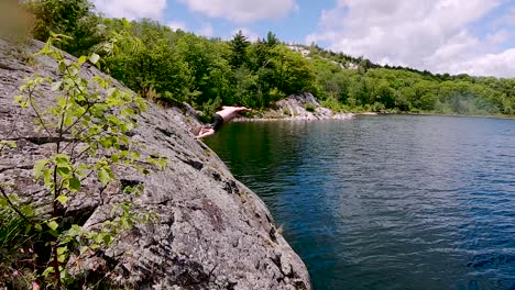 young white man jumps off rock into blue lake in slow motion, wide pan