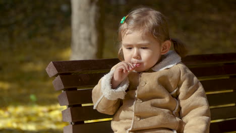 adorable blonde preschooler girl enjoy licking lollipop sitting on banch in autumn park in slow motion at sunset