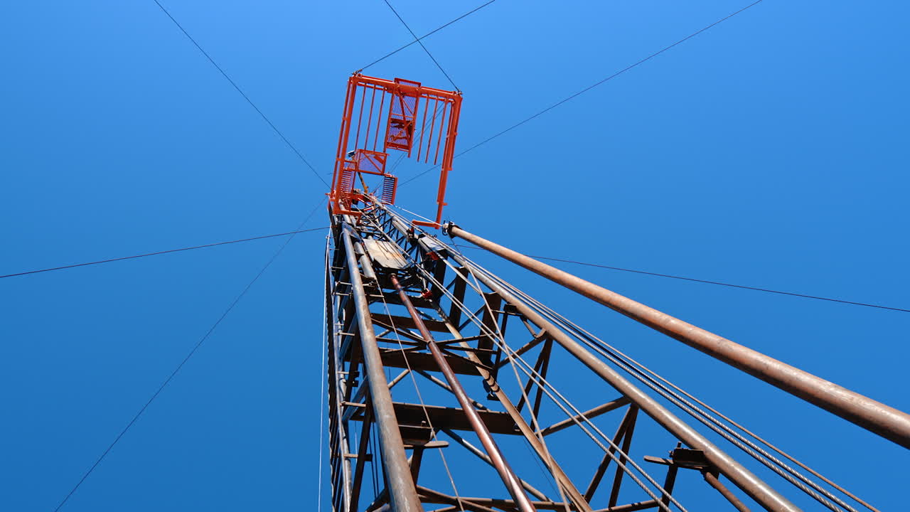 Looking up at the high metal tower with the wires to the sides attached to it. Low angle view on the industrial equipment for oil or gas production.