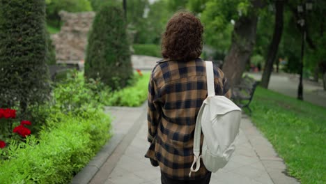 Back-view-A-brunette-girl-in-a-checkered-shirt-with-a-white-backpack-walks-through-the-park-after-classes-at-the-university