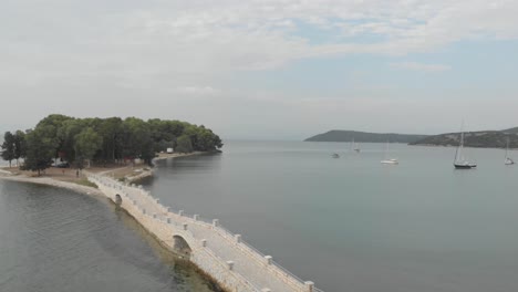 aerial drone footage with two people on bridge road arriving at small greek exotic island with sailing boats on the background , with  koukoumitsa vonitsa