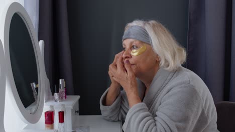 old senior elderly woman grandmother applying anti-wrinkle eye patch, putting makeup on at home