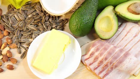 A variety of healthy fats including almonds, sunflower seeds, avocado, butter, and bacon on a wooden surface with bright lighting