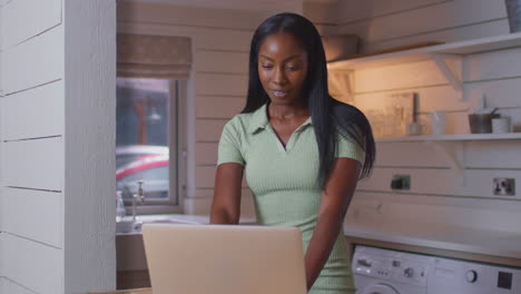 mujer joven de pie en el mostrador de la cocina con una computadora portátil trabajando desde casa