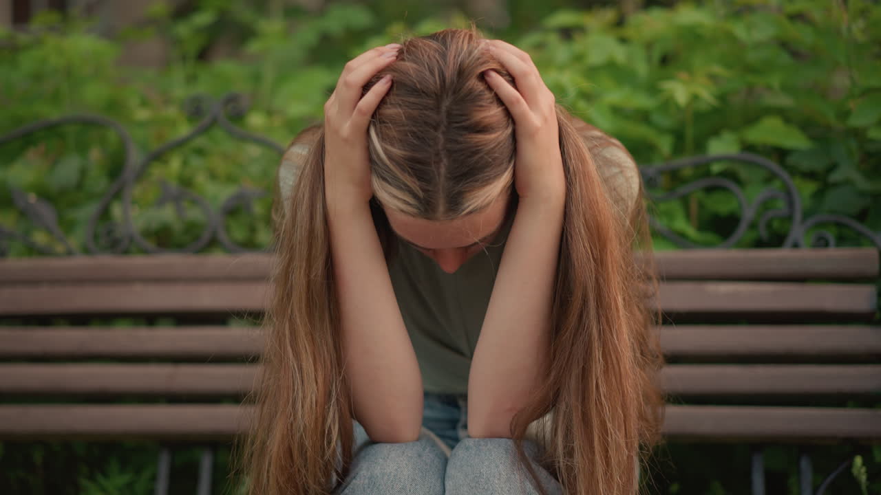 나무 벤치에 앉아있는 여자가 앞으로 구부러져 머리를 손에 고, 긴 머리카락이 아래로 떨어지며, 그녀는 초록색과 나무의 배경에서 사려 깊고 내성적으로 보입니다.