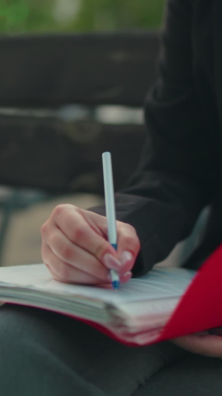 Close up of lady in professional attire flipping through red folder with pen in hand, seated next to laptop outdoors, with blurred bench and greenery in background