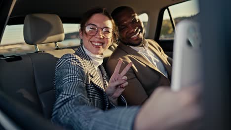 A-happy-brunette-girl-in-round-glasses-takes-a-selfie-with-her-colleague-a-Black-skinned-businessman-in-a-brown-suit-during-their-shared-trip-in-the-interior-of-a-modern-car