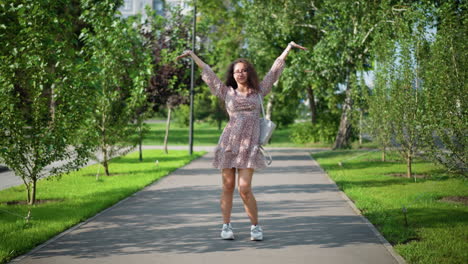 mujer alegre en vestido vintage bailando al aire libre con las manos levantadas, emanando felicidad contra un telón de fondo verde exuberante con edificios lejanos, el camino iluminado por el sol añade energía vibrante