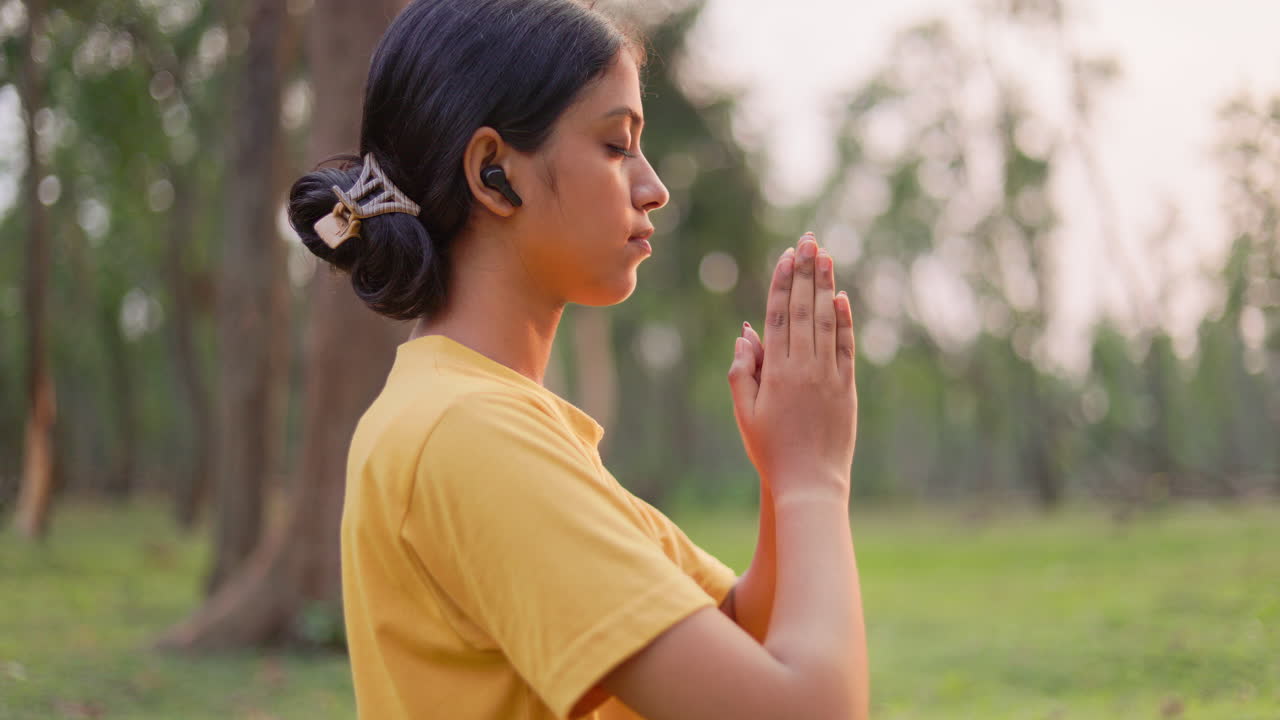 Donna serena che esegue il gesto della mano Namaste con gli auricolari wireless, dedicandosi alla meditazione e alla tranquillità nel verde durante l'ora d'oro