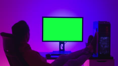 joven freelance descansando en una silla, bebiendo café y mirando un monitor de computadora con una llave de croma de pantalla verde, adentro por la noche en una habitación llena de luz de neón.