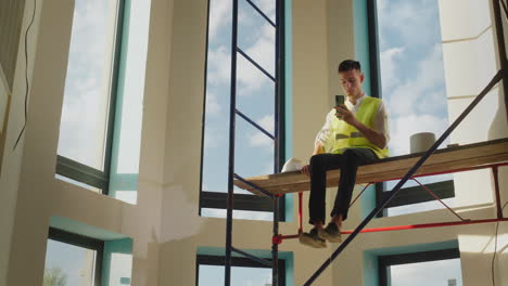 a bedless worker sits on scaffolding without a helmet, playing on a smartphone. dangerous behavior concept