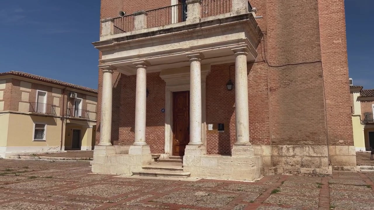 Panoramica della facciata d'ingresso della Cappella Reale di San Isidro a Cortijo de San Isidro, che evidenzia colonne in pietra calcarea, un balcone superiore, mattoni storici e le case tradizionali circostanti