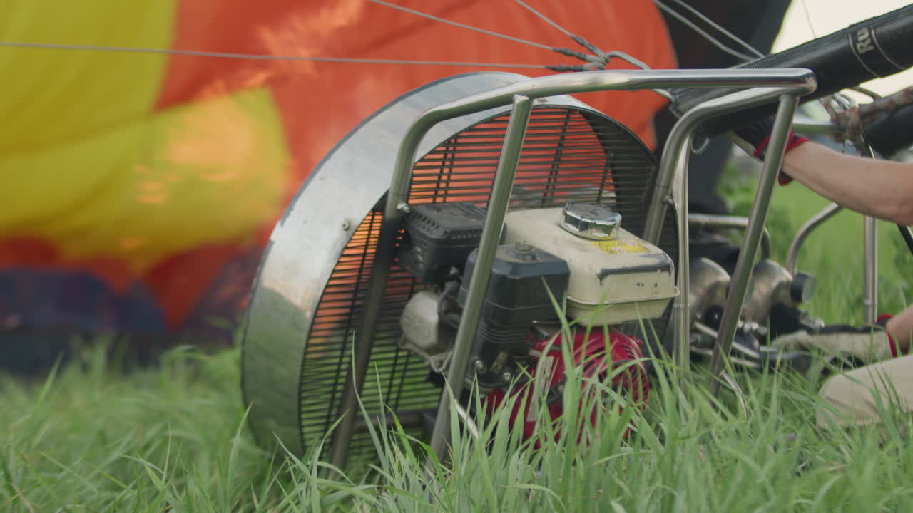 Person preparing hot air balloon for lift off as engine fan powers up and burner ignites fire beside vibrant deflated envelope resting on grass field during windy outdoor setup process