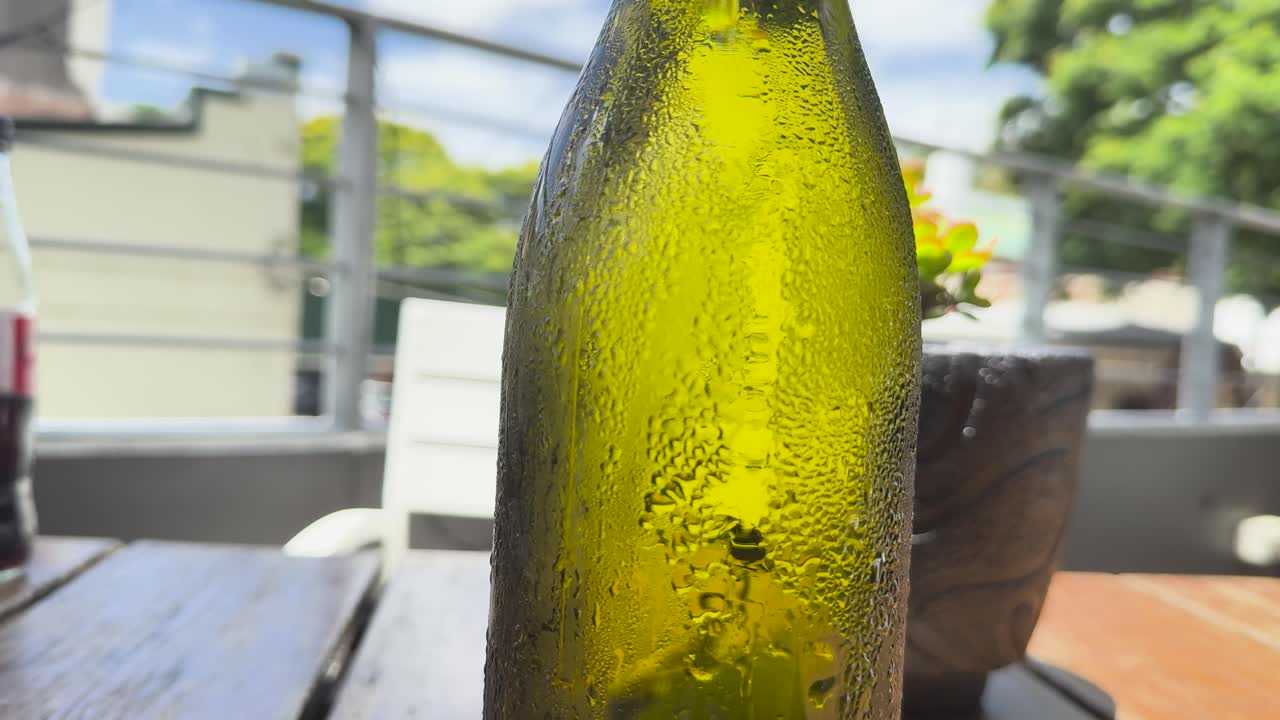 A green bottle with condensation is touched outdoors, highlighting moisture and sunlight interaction on a table