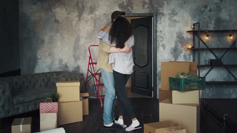 interracial friends relocating into new house. man raising woman, hugging and circling her in room with cardboard boxes, sofa, shelving and packages