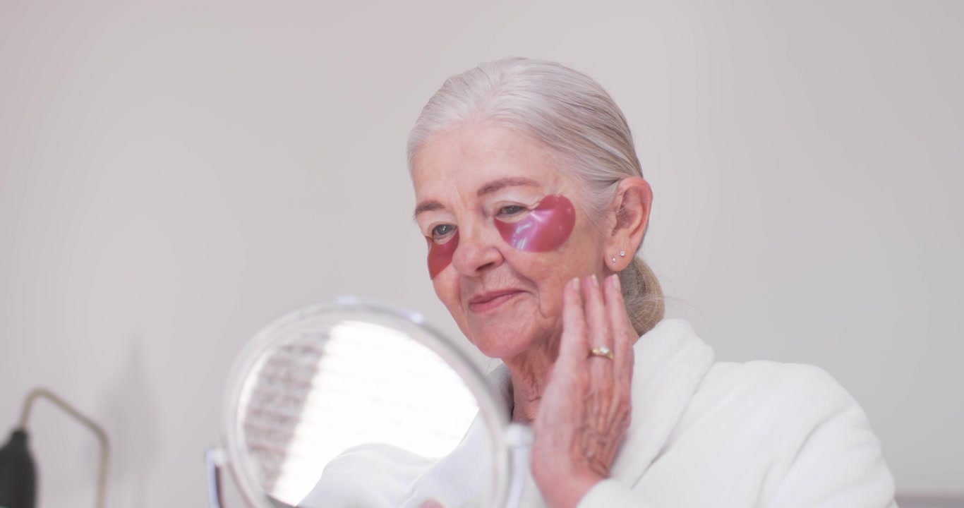 Con cerotti a forma di cuore rosa, donna anziana seduta alla toeletta in accappatoio, tiene uno specchio e regola i cerotti.