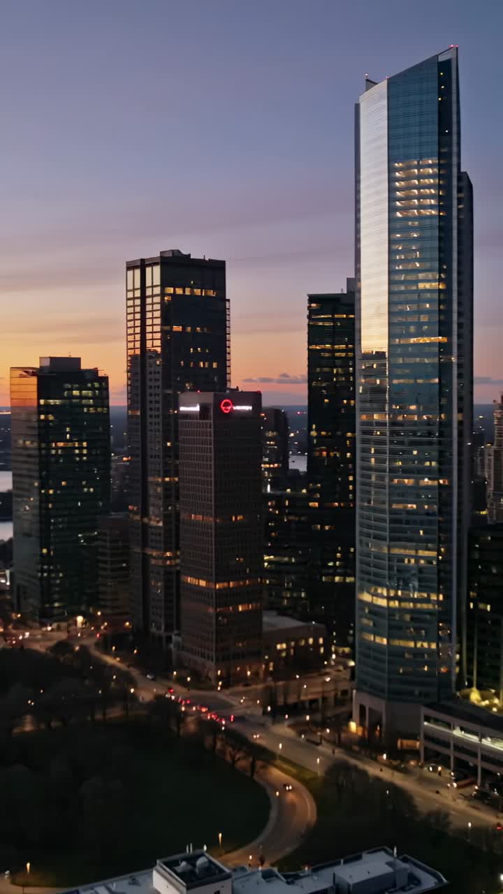 Vertical video: Dusk signaling drone panning urban skyline, revealing glass towers roadway by plaza