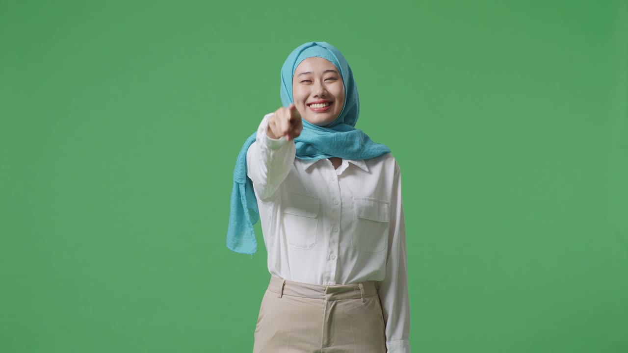 아시아의 무슬림 여성이 웃으며 가을 만지며 녹색 스크린 배경 스튜디오에 서있는 동안 당신을 가리키고 있습니다.