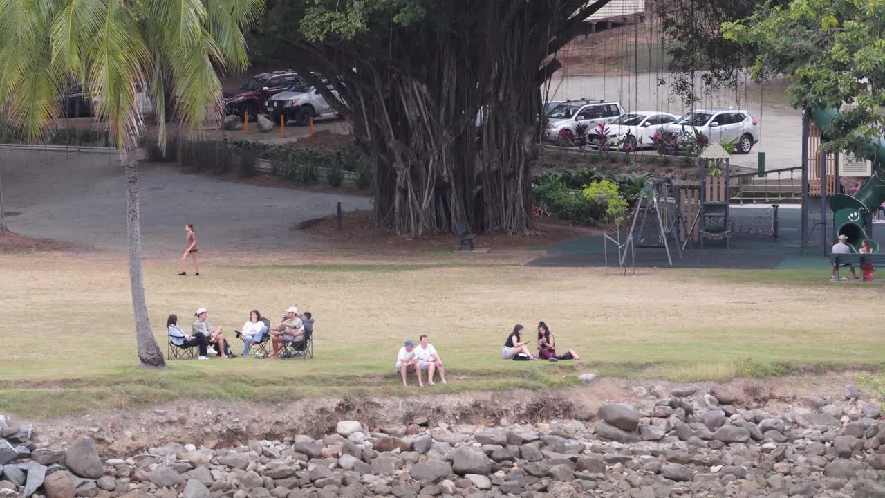 A serene picnic scene with people relaxing by a river under large trees in Port Douglas, captured in natural daylight